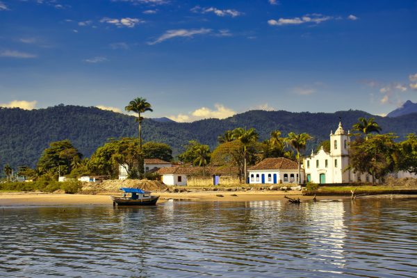 Barco no mar na frente de uma igreja antiga, com árvores e montanhas ao fundo, na cidade de Paraty